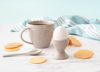 Boiled egg in ceramic egg cup, cup of coffee and thin crispy corn chips on background of turquoise kitchen towel. Breakfast concept. Copy space.