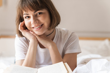 Fototapeta premium Happy cheerful positive young beautiful woman indoors at home lies on bed reading book.