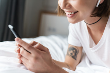 Positive young beautiful woman lies indoors at home on bed listening music with bluetooth earphones...