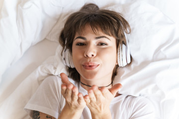 Pleased young beautiful woman lies indoors at home on bed listening music with headphones blowing kisses.
