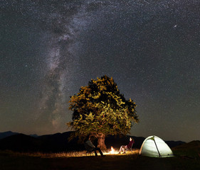 Romantic couple hikers enjoying summer night camping in mountains. Man and woman sitting on chairs beside bonfire, glowing tourist tent and big tree under starry sky full of stars and Milky way.