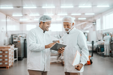 Young Caucasian supervisor showing results of food quality on tablet to his older colleague. Senior man holding folder with charts. Both are dressed in uniforms and having hairnets. Food plant.