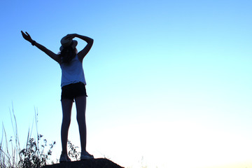 Silhouette of a young teenage girl  wearing a hat standing back, looking to  the sunset and holding hands up. People outdoors, travel, landscape concept. 