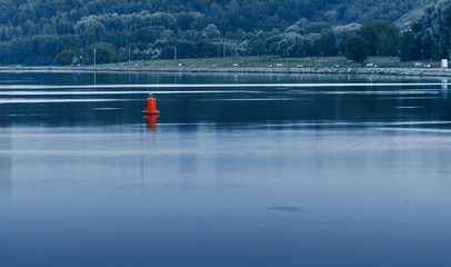 The red buoys of cylindrical shape in an urban river with calm water against the embankment with woodland Park.