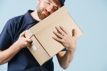 Cheerful delivery man in uniform is hugging cardboard box with his eyes closed