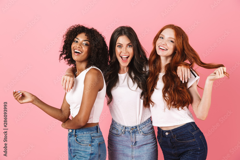 Wall mural Cheery smiling positive young three multiethnic girls friends posing isolated over pink wall background. - Wall murals