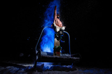 Active male snowboarder dressed in a white and pink sportswear sitting on the funicular seat rising hand