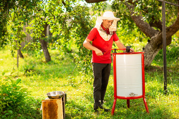 A beekeeper in a protective cap launches into work a honey extractor on the bee farm. Beekeeping