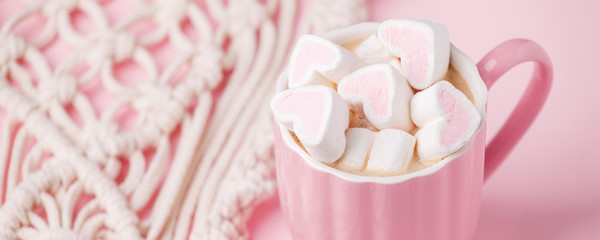 Cup of hot chocolate with pink heart shaped marshmallows and macrame panel on pink background,...