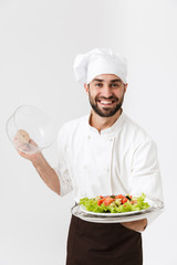Image of happy chief man in cook uniform smiling and holding plate with vegetable salad