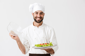 Image of joyful chief man in cook uniform smiling and holding plate with vegetable salad isolated over white wall