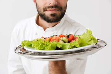 Cropped image of young chief man in cook uniform smelling dish while holding plate with vegetable salad