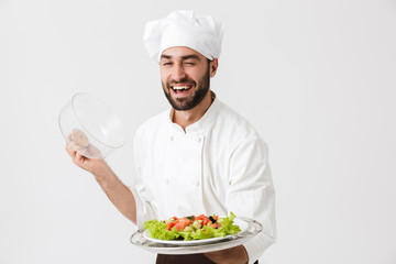 Image of handsome chief man in cook uniform smiling and holding plate with vegetable salad
