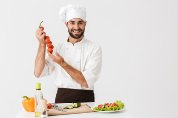 Image of caucasian chief man in uniform holding tomatoes while cooking vegetable salad on wooden cutting board