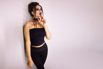 young stylish sexy brunette girl with halloween bloody makeup is standing and looking straight with fingers near her tongue in open mouth on white wall background, halloween concept, free space