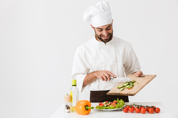 Image of positive cook man in uniform smiling and cutting vegetable salad on wooden board