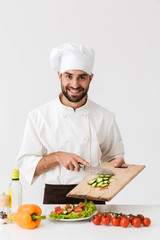 Image of handsome cook man in uniform smiling and cutting vegetable salad on wooden board