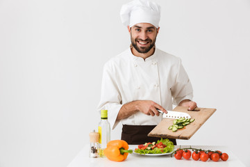Image of professional cook man in uniform smiling and cutting vegetable salad on wooden board