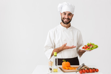 Image of positive cook man in uniform smiling and holding plate with vegetable salad