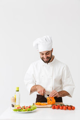 Image of pleased cook man in uniform smiling and cutting vegetable salad on wooden board