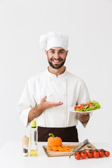 Image of caucasian cook man in uniform smiling and holding plate with vegetable salad