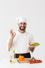 Image of young cook man in uniform showing ok sign and holding plate with vegetable salad