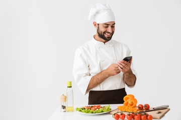 Image of pleased chef man in uniform smiling and holding smartphone while cooking vegetable salad