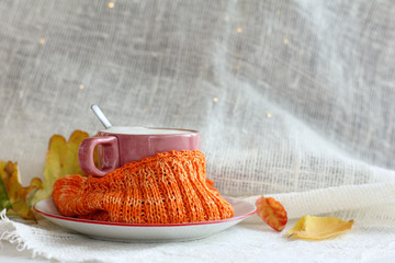 coffee mug wrapped in an orange scarf on a table next to autumn leaves. warming still life with cappuccino