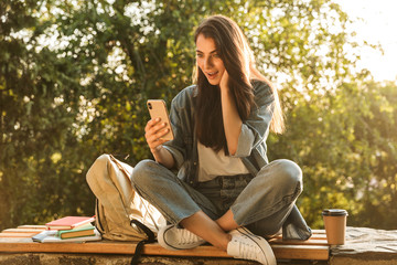 Image of happy brunette woman smiling and using smartphone while sitting at bench in green park