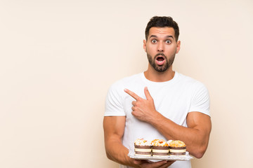 Handsome man holding muffin cake over isolated background surprised and pointing side