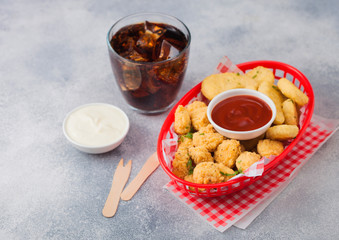 Buttered chicken nuggets and popcorn bites in red fast food basket with ketchup and glass of cola on light background.
