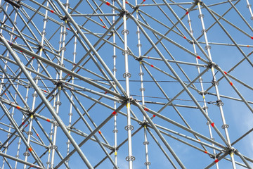 Fototapeta premium Metal pipe frame structure on blue sky background. Industrial construction from tubes raises upwards. Building process and joining elements view. Straight and inclined lines backdrop