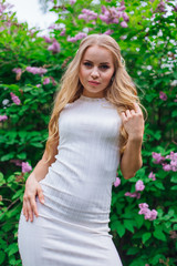 Portrait of a charming blond woman wearing beautiful white dress standing next to lilac bush.