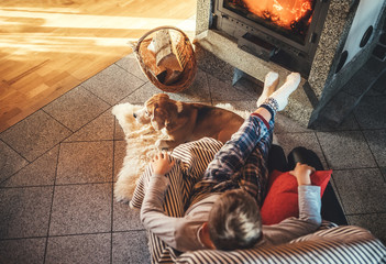 Boy sitting in comfortable armchair in cozy country house near fireplace and enjoying a warm atmosphere and flame moves. His beagle friend dog lying beside on the white sheepskin.