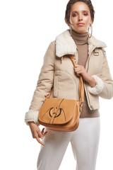 Medium full shot of a young chestnut European lady with a shoulder bag is wearing white casual trousers, a beige turtleneck and a short cream jacket with a fur trim. The bag is made of smooth sandy  © RedUmbrella&Donkey
