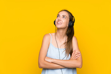 Young woman listening music over isolated yellow wall looking up while smiling