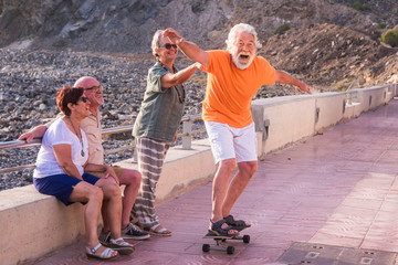 group of seniors and mature people at the beach have fun looking at old man riding a skateboard and...