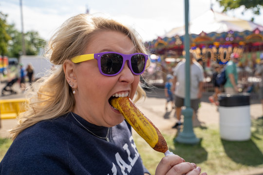 Blonde woman eats a pronto pup corn dog covered in mustard at an outdoor fair