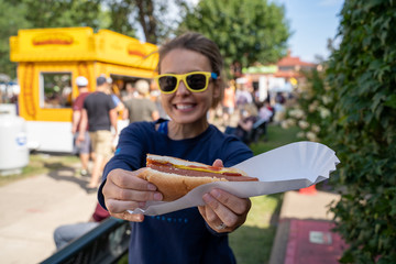 Smiling woman at an outdoor fair holds up a half-eaten footlong hot dog. Focus on the hot dog, girl...