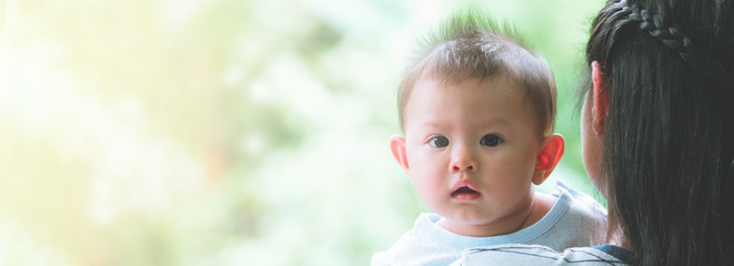 Young asian mother carrying her male toddler with green trees and garden in background. Toddler...
