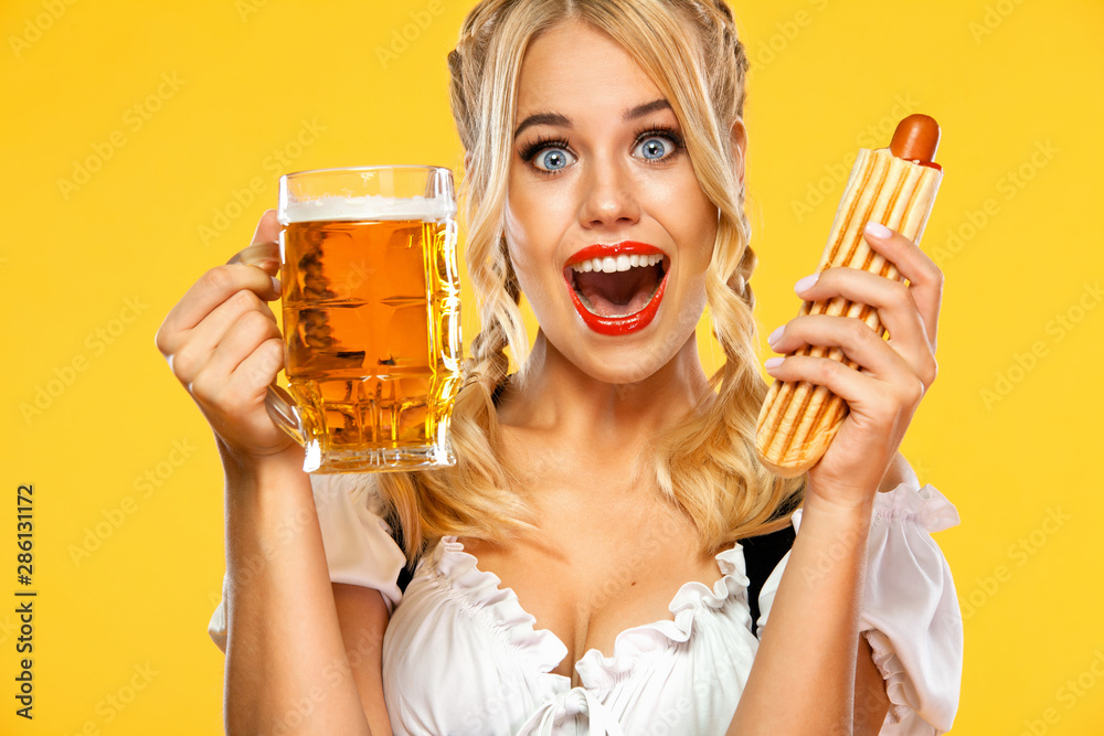 Wall mural Young sexy Oktoberfest girl waitress, wearing a traditional Bavarian or german dirndl, serving big beer mugs with drink and french hot dog isolated on yellow background.