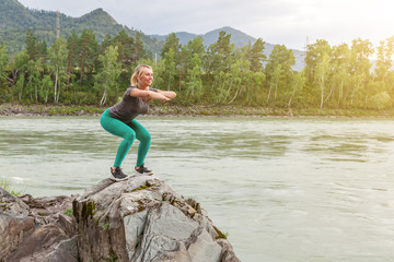 Blond pretty woman with sexy tight booty is doing stretching by squats stretching arms forward on the cliff in picturesque place in mountains near the river  and tree on stones. Work out and athletics