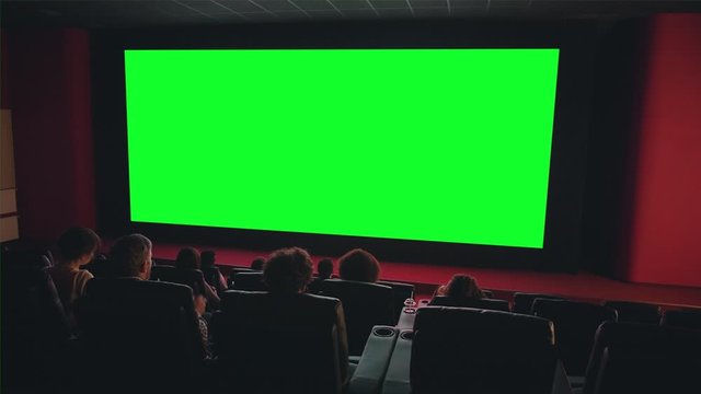 People audience are looking at big green chroma key screen in cinema enjoying film indoors in dark room. Entertainment, leisure and modern technology aoncept.