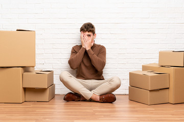 Handsome young man moving in new home among boxes covering eyes and looking through fingers