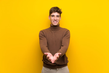 Handsome young man over isolated yellow background holding copyspace imaginary on the palm to...