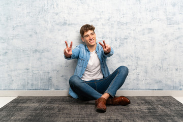 Handsome young man sitting on the floor smiling and showing victory sign