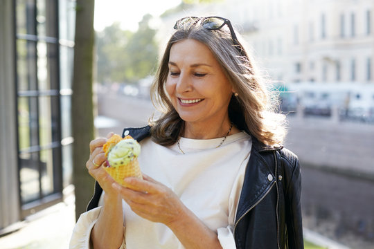 People, age, modern lifestyle, summertime and food concept. Attractive cheerful middle aged Caucasian woman wearing trendy black leather jacket smiling broadly, eating ice cream cone on street