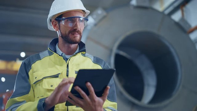 Portrait of Professional Heavy Industry Engineer / Worker Wearing Safety Uniform and Hard Hat Uses Tablet Computer. In the Background Construction Factory for Oil, Gas and Fuels Transport Pipeline