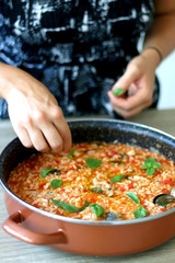 Unrecognizable person putting basil leaves in pot of orzo pasta, with tomato sauce, vegetables and seafood. Selective focus.