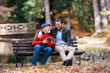 Senior father and his son sitting on bench by lake in nature, using tablet.